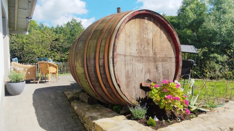 Een groot houten vat staat in de tuin, omringd door kleurrijke bloemen. Op de achtergrond zijn comfortabele tuinmeubelen en bomen te zien.