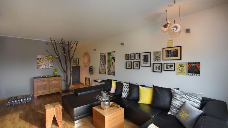 Un salon moderne avec un grand canapé noir et des œuvres d'art aux murs. Il y a une armoire en bois et une table en bois ainsi qu'un éclairage élégant.