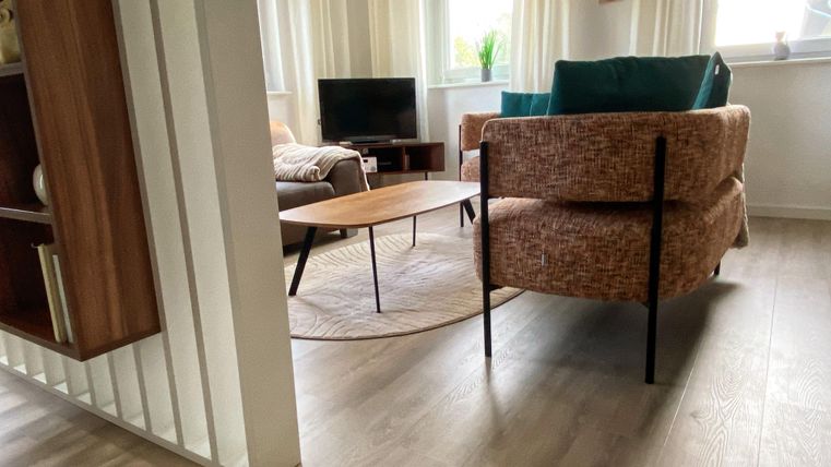 Ein modernes Wohnzimmer mit einem gemütlichen Sessel und einem Holzkaffee-Tisch. Große Fenster sorgen für viel Tageslicht und eine angenehme Atmosphäre.