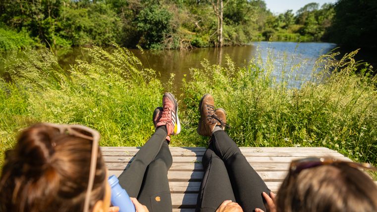 Zwei Personen entspannen auf einer Bank am Ufer eines Teiches, umgeben von grüner Natur.