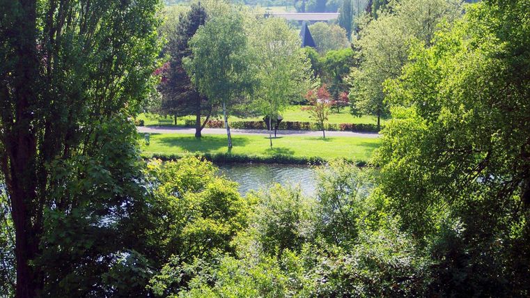 Ein ruhiger Fluss fließt durch eine grüne Landschaft mit vielen Bäumen. Im Hintergrund sind sanfte Hügel zu sehen, die eine friedliche Atmosphäre schaffen.