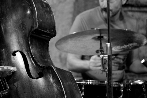 Ein Kontrabass steht im Vordergrund, während ein Schlagzeuger im Hintergrund spielt. Die Szene vermittelt eine lebendige Jazz-Atmosphäre.