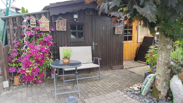 Ein gemütlicher Außenbereich mit einem Holzschuppen, Sitzgelegenheit und bunten Blumen. Der Raum ist freundlich gestaltet und lädt zum Entspannen ein.