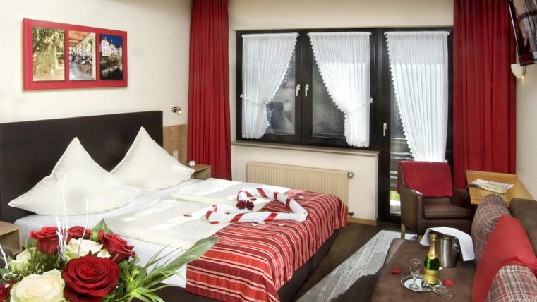 Une chambre d'hôtel confortable avec un grand lit et des rideaux rouges. Sur la table de nuit se trouvent des fleurs et il y a une bouteille de champagne prête.