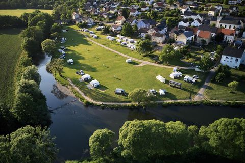 Een camping aan de rivier met veel campers en tenten. Op de achtergrond zijn huizen en een groene omgeving te zien.