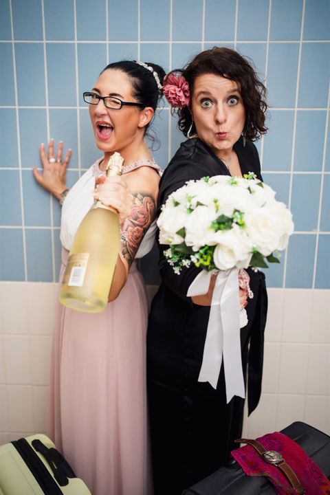 Zwei Frauen stehen fröhlich zusammen, eine mit Sektflasche und die andere mit einem Blumenstrauß. Sie feiern offensichtlich einen besonderen Anlass.