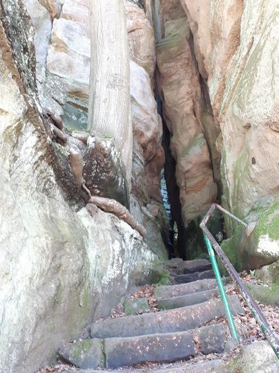 Un étroit chemin entre des rochers hauts mène à un escalier. Les pierres sont recouvertes de mousse et l'environnement semble naturel.
