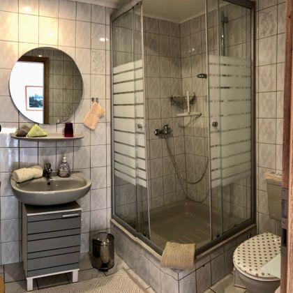 A modern bathroom with a shower and a sink. The walls are tiled in white, and there is a round mirror as well as a toilet.