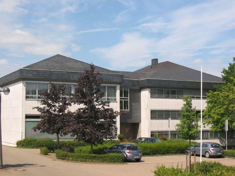 Een modern gebouw met een hellend dak en grote ramen. Omringd door bomen en enkele geparkeerde auto's.