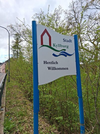 Un panneau de bienvenue de la ville de Kyllburg. Le panneau montre un paysage vert avec des arbres en arrière-plan.