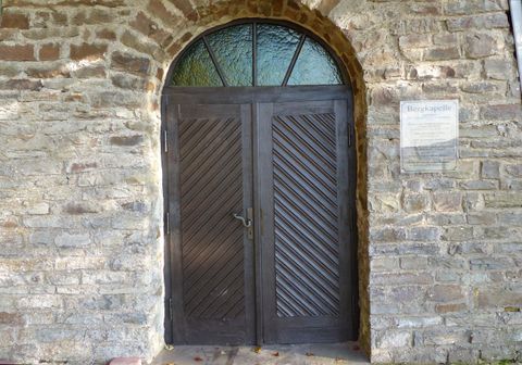 Eine dunkle Holztür mit einem bogenförmigen Oberlicht. Die Tür ist in eine Steinmauer eingelassen und hat diagonale Muster.