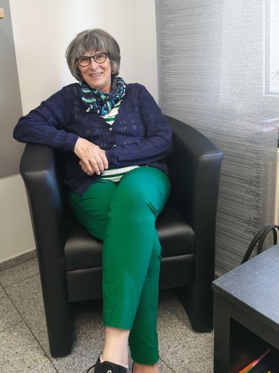 Eine lächelnde Frau sitzt in einem schwarzen Sessel. Sie trägt einen blauen Pullover und grüne Hosen.