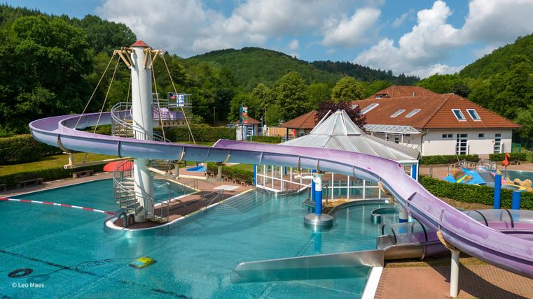 Ein Schwimmbad mit einer langen, lilaRutsche und klarem Wasser. Im Hintergrund sind Bäume und ein Gebäude zu sehen.