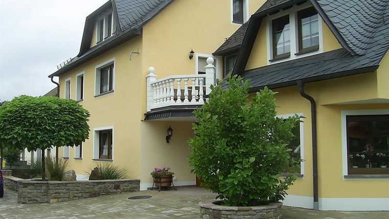 Ein modernes Haus mit gelber Fassade und einem Balkon. Vor dem Haus befinden sich Bäume und eine Pflastersteinfläche.