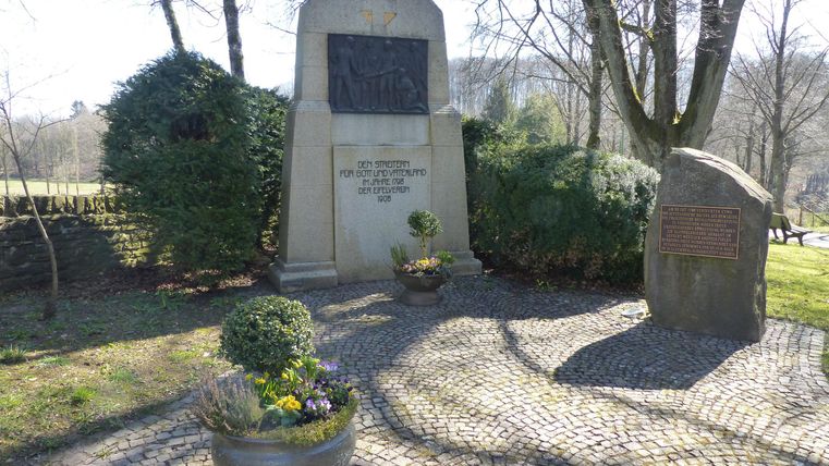 Ein Denkmal in einer ruhigen Umgebung mit Bäumen und Sträuchern. Der Bereich ist mit Kieselsteinen ausgelegt und enthält Blumengefäße.