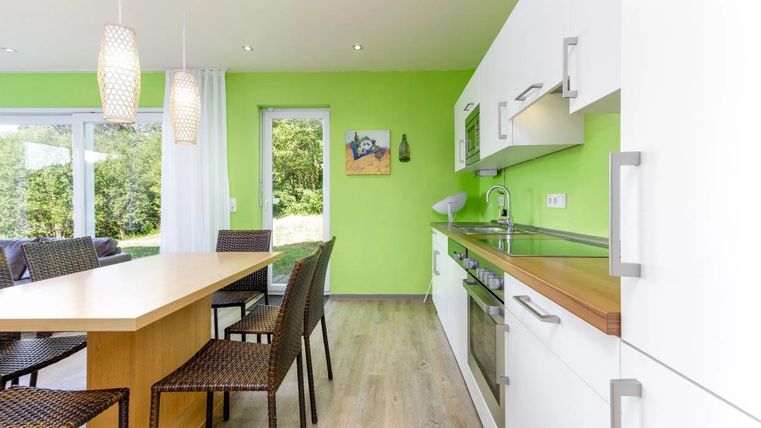Een moderne keuken met groene muren en lichte meubels. De eethoek bestaat uit een grote tafel en stoelen van riet.