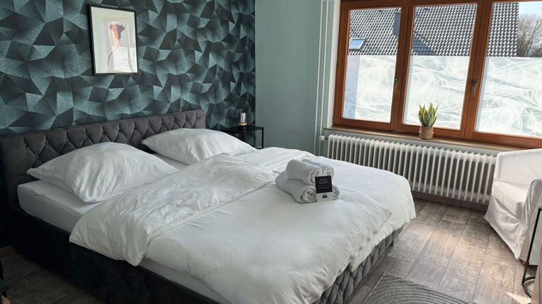Ein modernes Schlafzimmer mit einem großen Doppelbett und einem geometrischen Wanddesign. Das Zimmer hat große Fenster, die natürliches Licht hereinlassen.