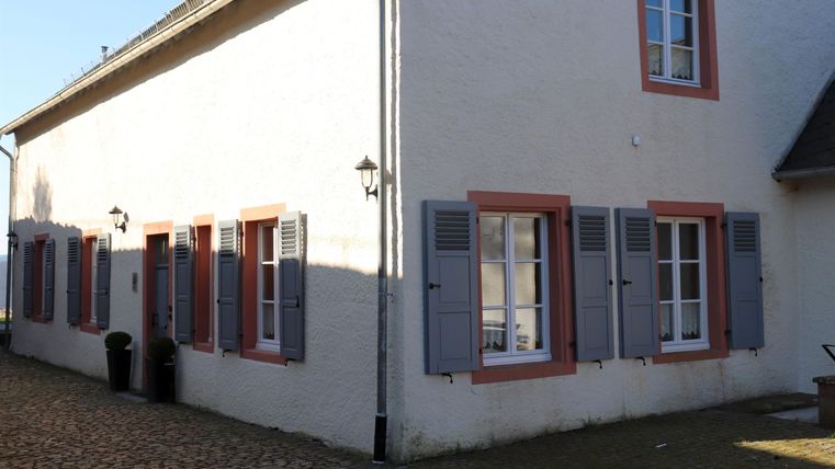 Ein charmantes Haus mit weißen Wänden und bunten Fensterläden. Die sonnige Atmosphäre und der gepflasterte Weg verleihen dem Ort einen einladenden Charakter.