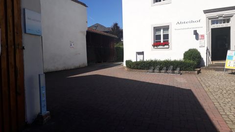 Ein heller Innenhof mit pavierten Wegen und weißen Wänden. Im Hintergrund ist ein Gebäude mit einem Fenster und grünen Sträuchern zu sehen.