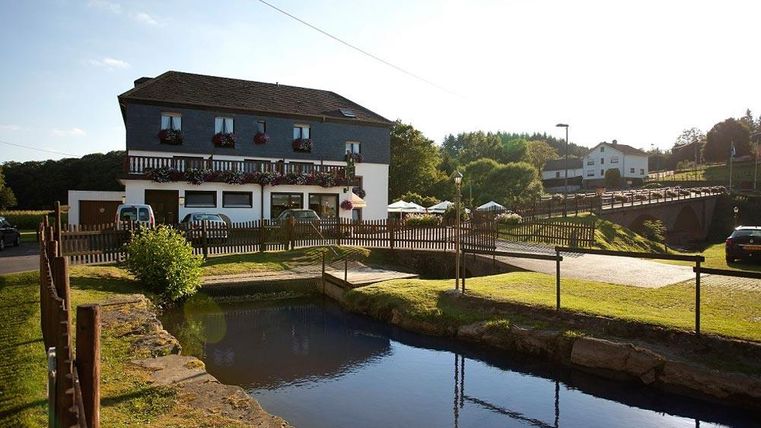 Une maison confortable avec des balcons et un étang paisible devant. À l'arrière-plan, on voit des prairies vertes et des arbres.