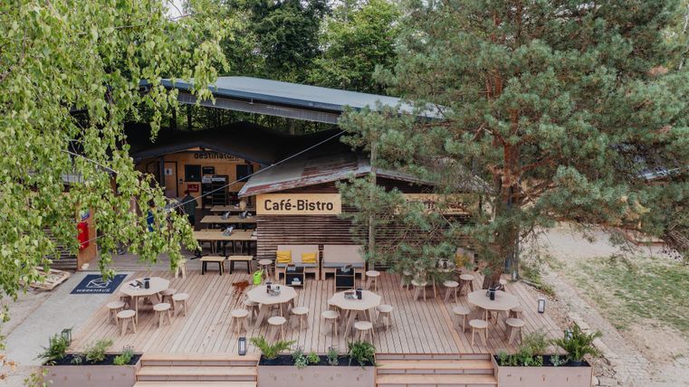 Ein gemütliches Café-Bistro mit einer Holzterrasse und runden Tischen. Umgeben von Bäumen bietet es einen einladenden Außenbereich.