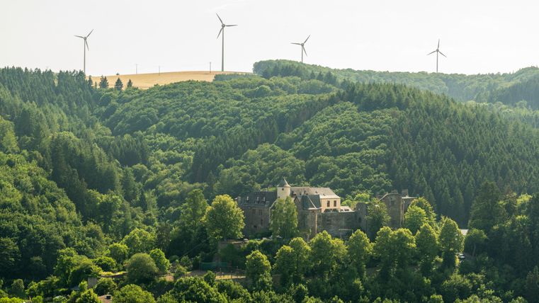 Eine malerische Landschaft mit sanften Hügeln und dichten Wäldern. Im Hintergrund sind Windräder zu sehen.