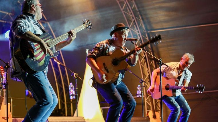 Drei Musiker spielen auf der Bühne Akustikgitarre. Die Atmosphäre ist lebhaft und festlich mit bunten Lichtern im Hintergrund.