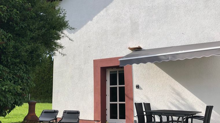 Une maison moderne avec une façade blanche et des bordures rouges. Sur la terrasse, il y a une table et des chaises ainsi que des transats dans le jardin.