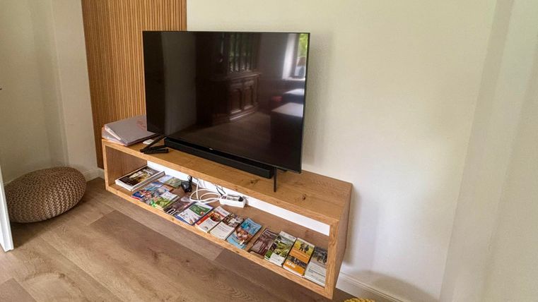 Ein moderner Fernseher auf einem Holzregal, das mit verschiedenen Zeitschriften gefüllt ist. Daneben befinden sich gemütliche Sitzkissen.