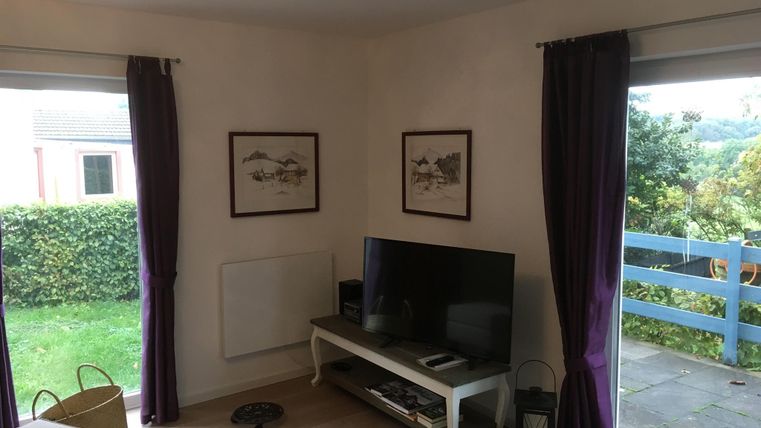 Ein gemütliches Zimmer mit einem Fernseher und dekorativen Bildern an der Wand. Durch die Fenster sieht man einen schönen Garten und einen blauen Balkon.