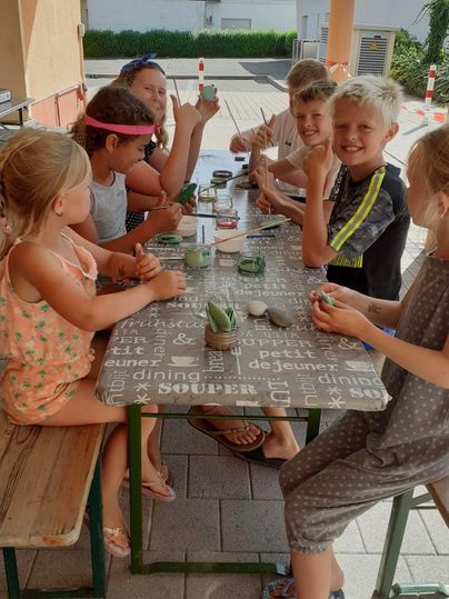 Un groupe d'enfants est assis à une table en plein air et crée des travaux manuels. Ils semblent joyeux et créatifs, tandis qu'ils présentent leurs projets.