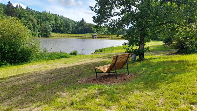 Eine ruhige Landschaft mit einem Teich und einem bequemen Stuhl am Ufer. Umgeben von Bäumen und Wiese bietet der Ort eine entspannte Atmosphäre.
