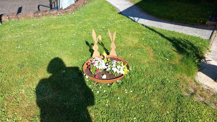 A round flower pot with colorful flowers and two wooden rabbit figures stands on the green lawn. In the background, a paved path is visible.