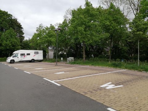 Een camper op een parkeerplaats met veel vrije staanplaatsen. De omgeving is groen met bomen.