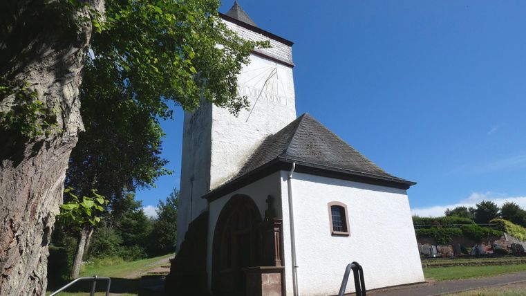 Eine alte Kirche mit einem weißen Turm und einem schattenspendenden Baum daneben. Der Himmel ist blau und klar.