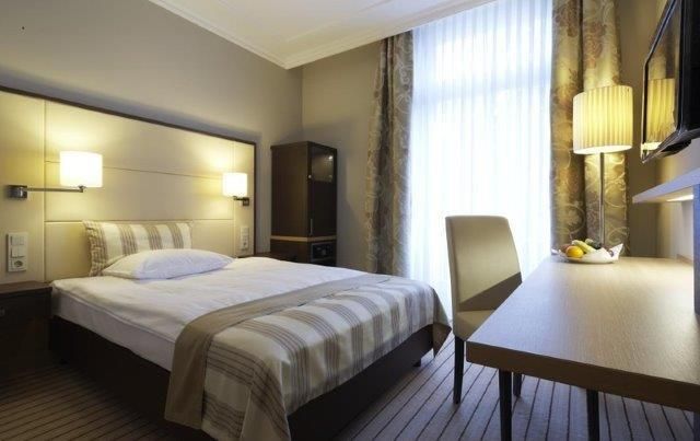 Een moderne hotelkamer met een tweepersoonsbed, een bureau en een televisie. De ramen laten veel daglicht binnen en zorgen voor een aangename sfeer.