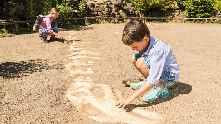 Twee kinderen spelen op een zandveld. Een jongen tekent iets in het zand, terwijl een meisje op de achtergrond kijkt.
