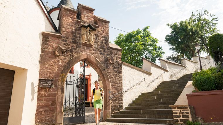 Ein historisches Tor aus Stein mit einem Wappen. Im Vordergrund läuft eine Frau in einem grünen Kleid an der Treppe vorbei.