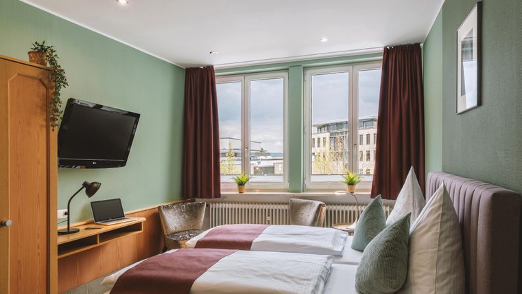 Ein gemütliches Zimmer mit zwei Einzelbetten und einem Schreibtisch. Große Fenster lassen viel Tageslicht herein und bieten einen Blick auf die Umgebung.