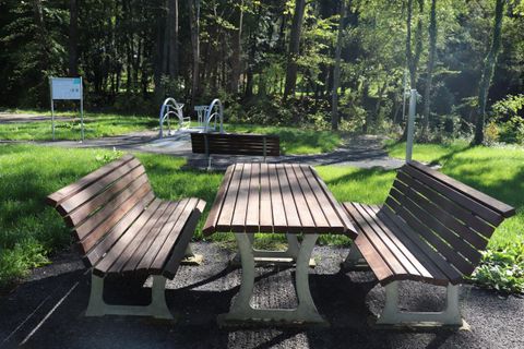 Une zone de repos pavée avec des bancs en bois et une table dans la forêt. En arrière-plan, on aperçoit des espaces verts et des équipements de jeu.