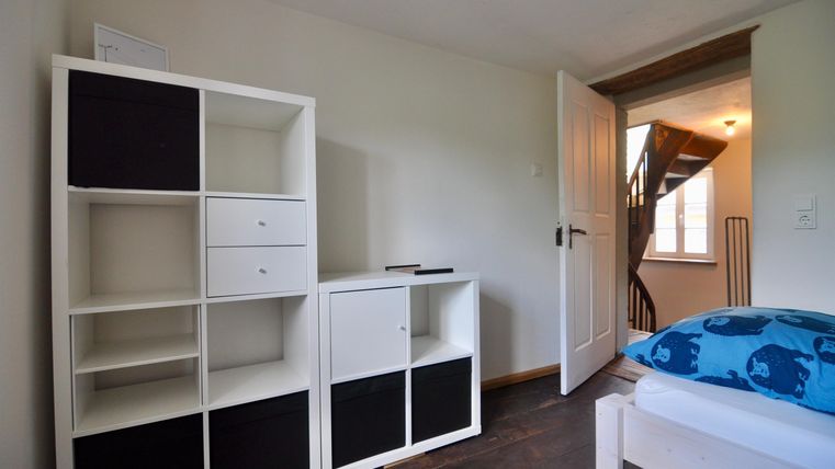 Ein einfaches Zimmer mit einem weißen Regal und einem Bett. Eine Tür führt zu einer Treppe im Hintergrund.