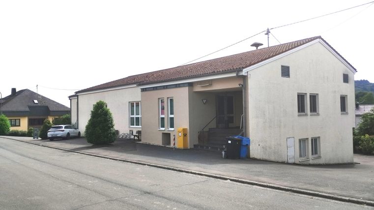 Ein einfaches Gebäude an einer Straße, umgeben von wenigen Bäumen und einem Parkplatz. Die Fassade ist hell und hat mehrere Fenster.