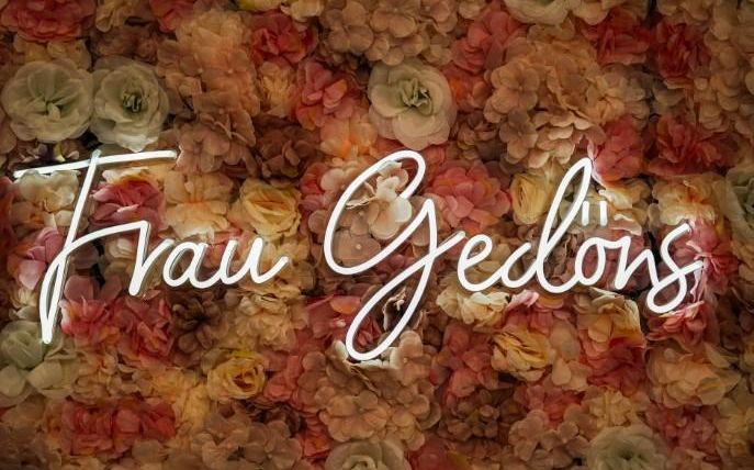 Ein Schriftzug "Frau Gedöns" vor einem Hintergrund aus bunten Blumen. Die Blüten sind in zarten Rosa- und Beigetönen gehalten.