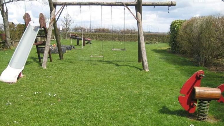 A playground with a slide and two swings is situated on a green meadow. Surrounded by bushes, it is ideal for children to play.