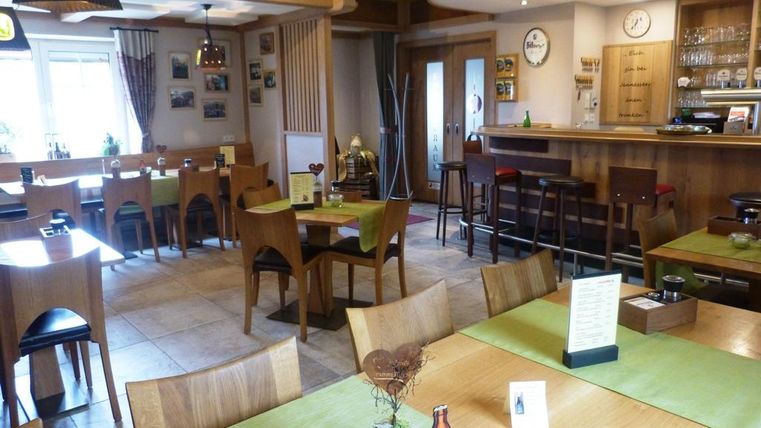 Ein gemütliches Restaurant mit Holzmöbeln und grünen Tischdecken. Die Wände sind mit Fotos dekoriert und eine Bar befindet sich im Hintergrund.