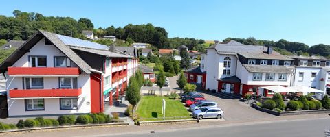 Ein modernes Hotelgebäude mit roten Akzenten und einem klaren Himmel. Die Umgebung ist grün und einladend, mit Autos auf dem Parkplatz.