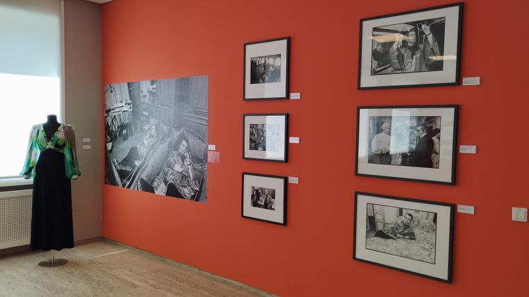 Ein Ausstellungsraum mit orangefarbene Wänden. An der Wand hängen Schwarz-Weiß-Fotografien und links steht ein Mannequin mit einem grünen Oberteil.