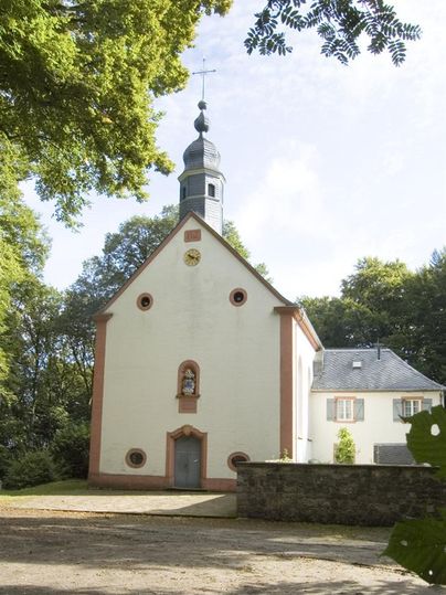 Une petite église charmante avec un clocher, entourée d'arbres. La façade est blanche avec des accents rouges et possède des fenêtres doubles accueillantes.