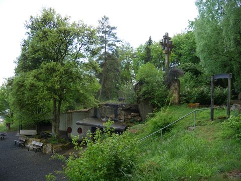 Eine malerische Landschaft mit vielen Bäumen und einer grünen Umgebung. Im Vordergrund sind Sitzbänke und einige Steinskulpturen sichtbar.
