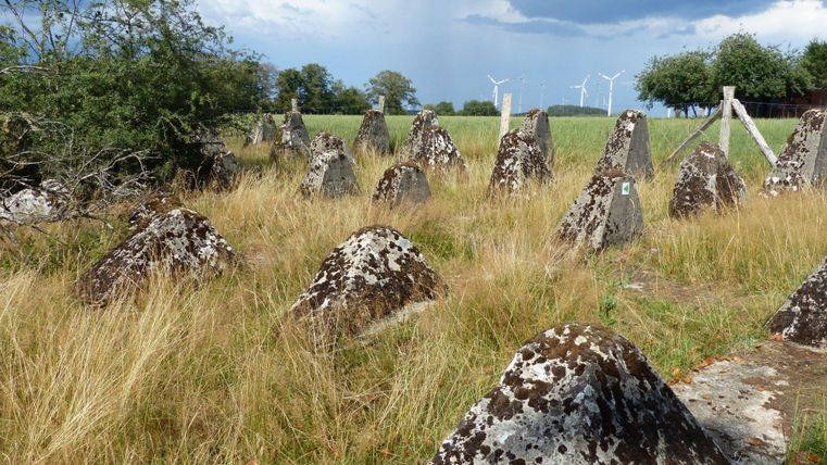 Eine Gruppe von steinernen Hügeln steht in einem offenen Feld. Im Hintergrund sind Windräder und ein bewölkter Himmel zu sehen.